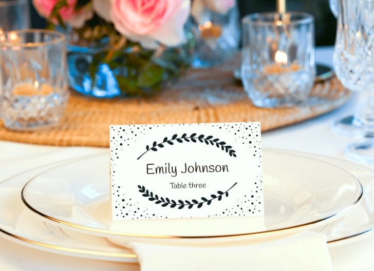 Egyszerű, mégis elegáns esküvői ültetőkártya fekete babér díszítéssel fehér tányéron, romantikus asztali beállítás része.